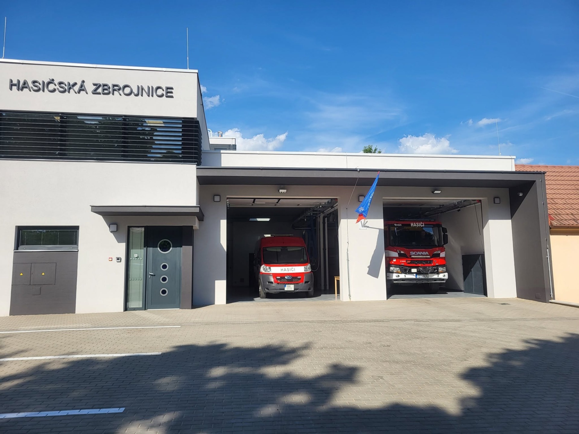 Před hasičskou zbrojnicí stojí červené hasičské vozy Scania, u vchodu socha a vlajky.